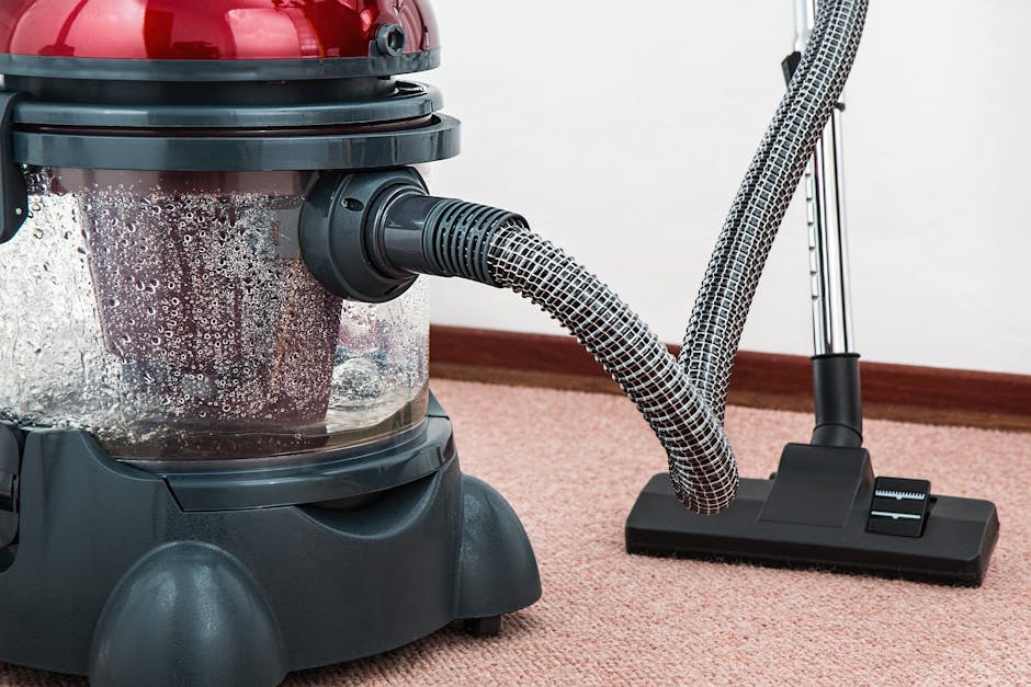 A person in a beige jumpsuit is performing surface cleaning in a modern indoor space, using a handheld vacuum cleaner connected to a larger, beige industrial vacuum unit on the floor. The room feature